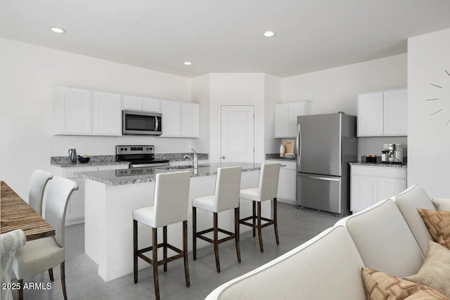 kitchen with stainless steel appliances, a kitchen island with sink, white cabinetry, light stone countertops, and a kitchen bar