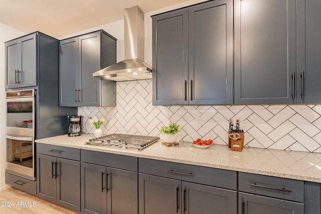 kitchen with light wood finished floors, tasteful backsplash, wall chimney exhaust hood, appliances with stainless steel finishes, and light stone counters