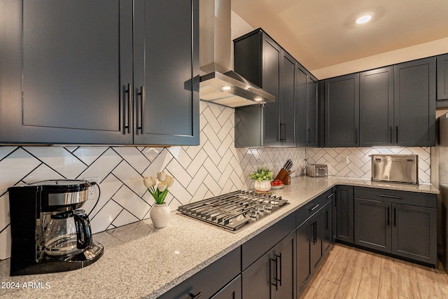 kitchen with decorative backsplash, wall chimney exhaust hood, light wood-style flooring, light stone counters, and stainless steel gas cooktop