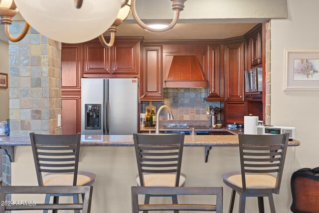 kitchen featuring stainless steel fridge, a kitchen breakfast bar, custom range hood, dark stone counters, and backsplash