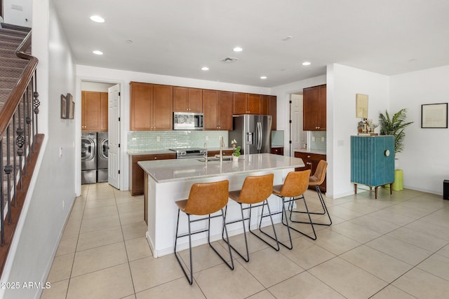 kitchen featuring an island with sink, backsplash, stainless steel appliances, separate washer and dryer, and light countertops