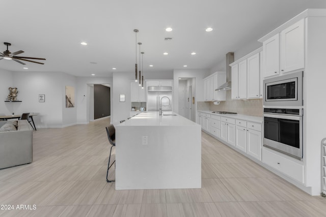 kitchen featuring pendant lighting, a center island with sink, white cabinets, wall chimney range hood, and stainless steel appliances