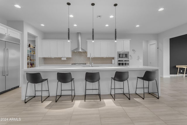 kitchen featuring decorative backsplash, wall chimney exhaust hood, a large island with sink, built in appliances, and decorative light fixtures