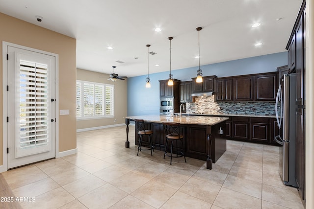 kitchen featuring pendant lighting, backsplash, light stone counters, stainless steel appliances, and a center island with sink