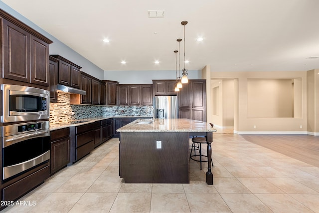 kitchen featuring stainless steel appliances, light stone counters, dark brown cabinetry, a center island with sink, and decorative backsplash