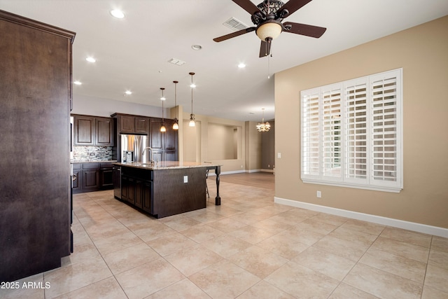 kitchen with a breakfast bar area, decorative light fixtures, dark brown cabinets, stainless steel fridge, and an island with sink