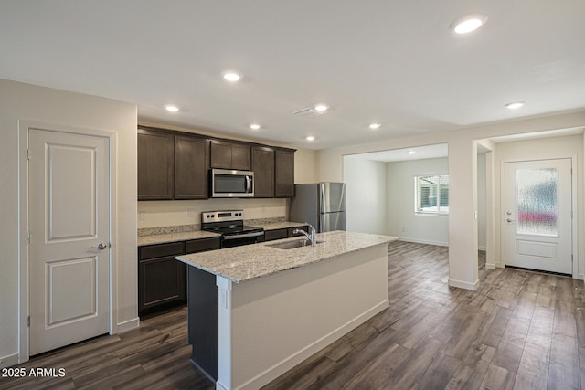 kitchen featuring a sink, appliances with stainless steel finishes, light stone countertops, dark wood finished floors, and a center island with sink