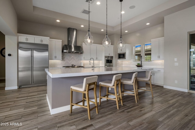 kitchen with visible vents, wood tiled floor, white cabinets, built in appliances, and wall chimney exhaust hood