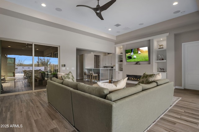 living area featuring visible vents, ceiling fan, built in features, wood finished floors, and a glass covered fireplace