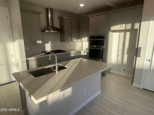 kitchen with wall chimney range hood, sink, stainless steel appliances, an island with sink, and decorative backsplash