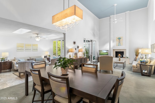 dining space with light colored carpet, ceiling fan with notable chandelier, a large fireplace, and a towering ceiling