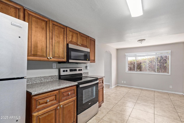 kitchen featuring dark countertops, baseboards, brown cabinets, arched walkways, and stainless steel appliances