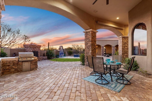 patio terrace at dusk featuring ceiling fan, a grill, and exterior kitchen