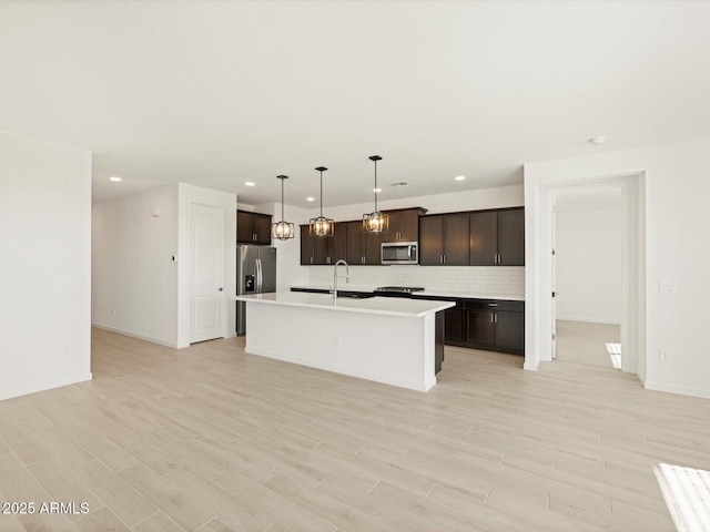 kitchen with appliances with stainless steel finishes, an island with sink, pendant lighting, dark brown cabinets, and backsplash