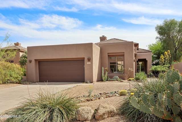 pueblo revival-style home featuring a chimney, driveway, stucco siding, and a garage
