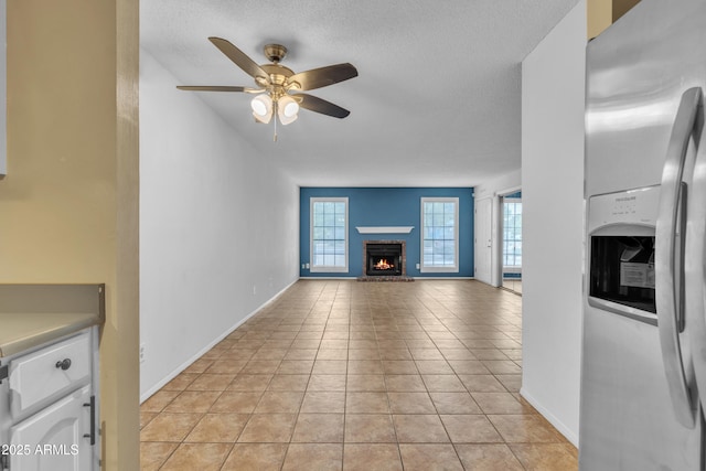 unfurnished living room with a brick fireplace, a ceiling fan, light tile patterned flooring, baseboards, and a textured ceiling