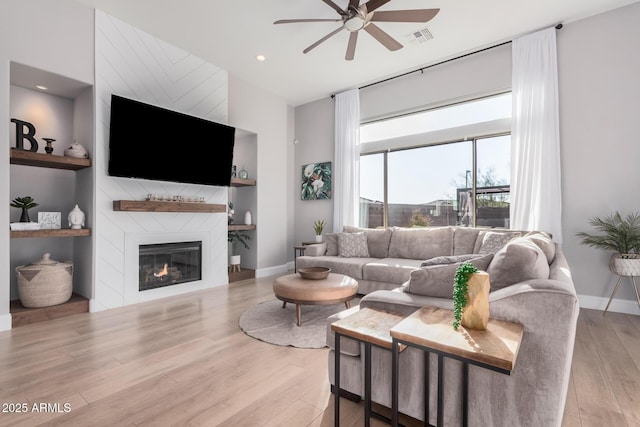 living room featuring a large fireplace, ceiling fan, light hardwood / wood-style floors, and built in shelves