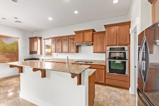 kitchen with appliances with stainless steel finishes, light stone counters, a center island with sink, light tile patterned floors, and a kitchen breakfast bar