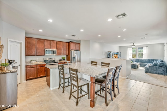 kitchen with a breakfast bar area, appliances with stainless steel finishes, light stone countertops, a center island with sink, and light colored carpet