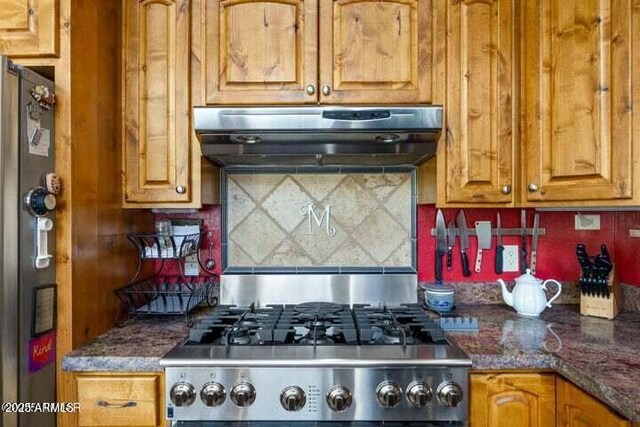 kitchen featuring dark stone countertops, exhaust hood, stainless steel fridge, stove, and backsplash