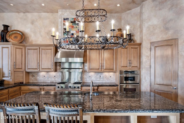 kitchen with double oven, an island with sink, pendant lighting, and wall chimney range hood