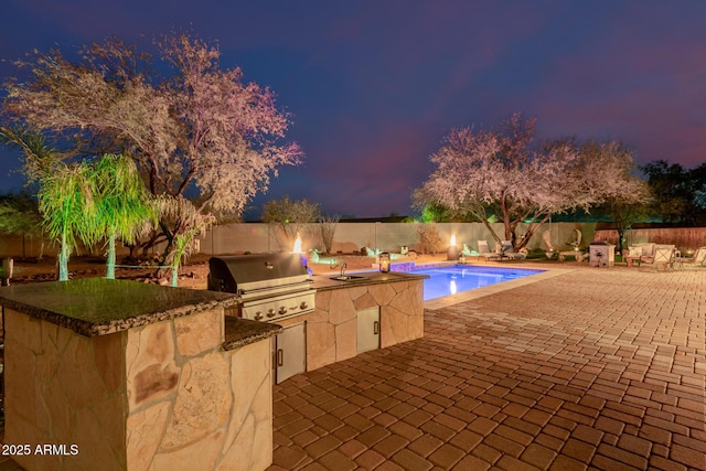 pool at dusk featuring exterior kitchen, an outdoor wet bar, grilling area, and a patio