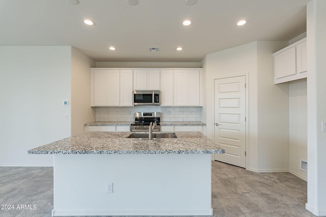 kitchen with light stone countertops, appliances with stainless steel finishes, backsplash, a center island with sink, and white cabinetry