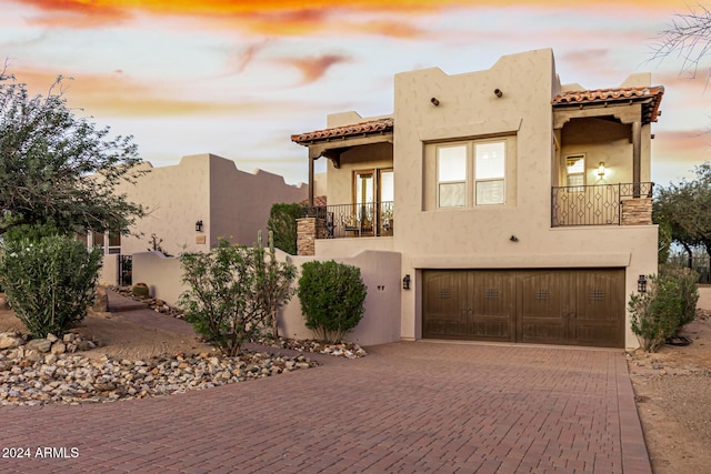 pueblo revival-style home featuring a tile roof, a garage, stucco siding, decorative driveway, and a balcony