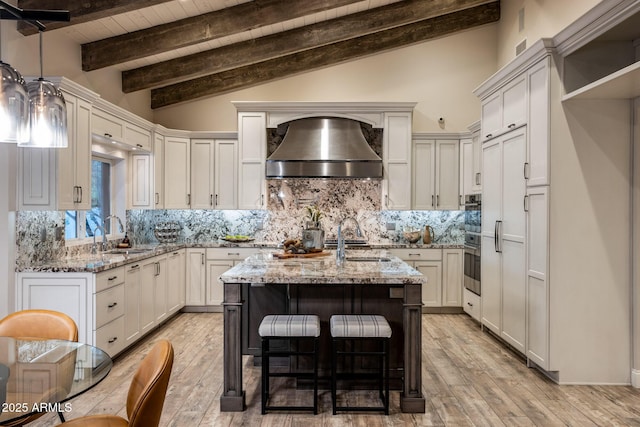 kitchen with sink, light stone counters, light hardwood / wood-style floors, a kitchen island with sink, and wall chimney range hood