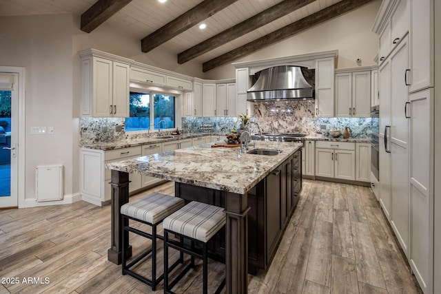 kitchen with a kitchen island with sink, lofted ceiling with beams, wall chimney exhaust hood, and light wood-type flooring