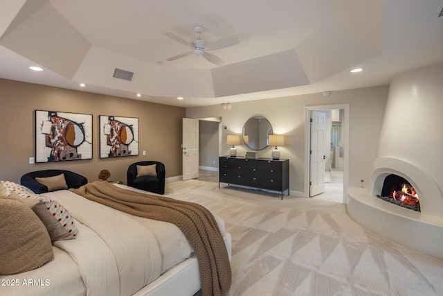 carpeted bedroom with ceiling fan, a raised ceiling, a large fireplace, and ensuite bath