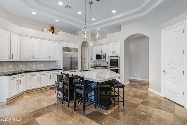 kitchen featuring appliances with stainless steel finishes, white cabinetry, backsplash, a center island with sink, and a raised ceiling