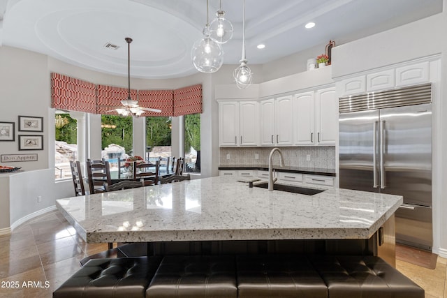 kitchen with sink, a kitchen island with sink, stainless steel built in refrigerator, a tray ceiling, and white cabinets