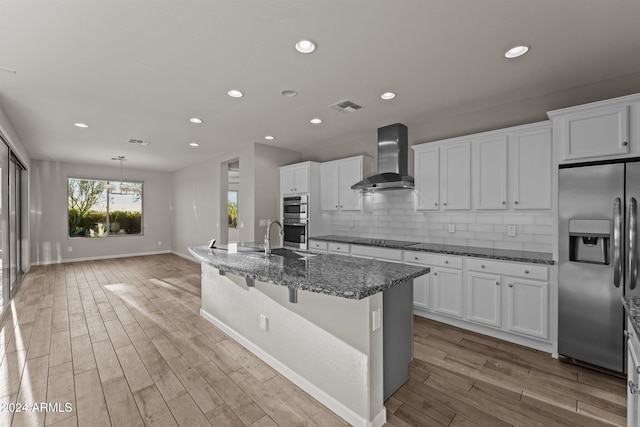 kitchen featuring wall chimney range hood, backsplash, light hardwood / wood-style floors, a center island with sink, and appliances with stainless steel finishes