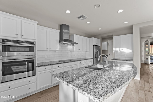 kitchen featuring wall chimney range hood, sink, an island with sink, light hardwood / wood-style floors, and stainless steel appliances