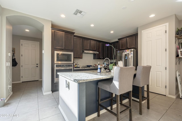 kitchen with stainless steel appliances, light stone countertops, a kitchen island with sink, and light tile patterned floors