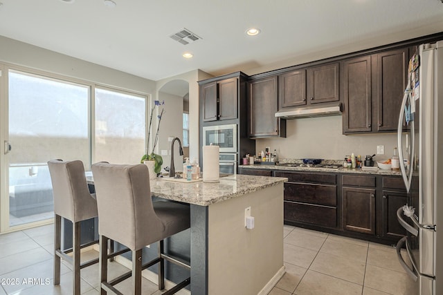 kitchen featuring a kitchen bar, light stone counters, a center island with sink, light tile patterned floors, and stainless steel appliances