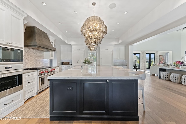 kitchen with premium range hood, hanging light fixtures, a large island with sink, light stone countertops, and range with two ovens