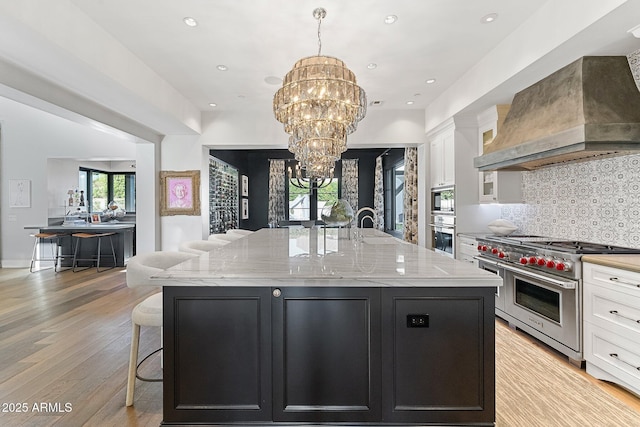 kitchen with white cabinetry, a center island with sink, double oven range, custom range hood, and light stone countertops