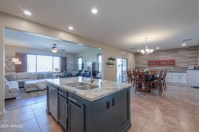 kitchen with light stone countertops, ceiling fan with notable chandelier, sink, dishwasher, and an island with sink