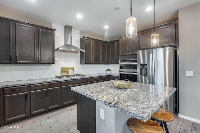 kitchen with wall chimney range hood, decorative backsplash, light wood-type flooring, appliances with stainless steel finishes, and decorative light fixtures