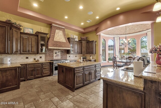 kitchen with a center island with sink, high end stainless steel range, an inviting chandelier, custom exhaust hood, and a sink