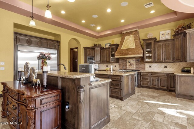 kitchen featuring a tray ceiling, a center island with sink, stainless steel appliances, visible vents, and premium range hood