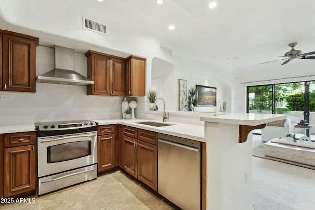 kitchen featuring visible vents, a peninsula, wall chimney exhaust hood, a sink, and stainless steel appliances