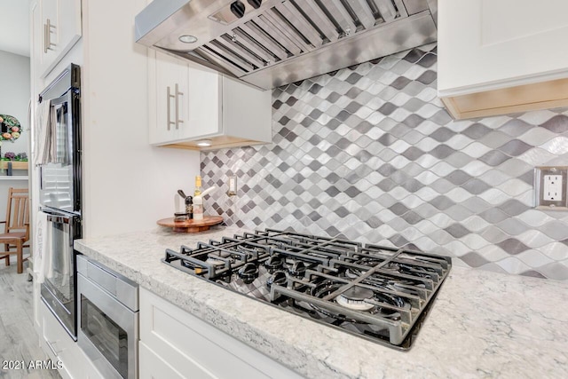 kitchen featuring tasteful backsplash, appliances with stainless steel finishes, light stone countertops, premium range hood, and white cabinetry