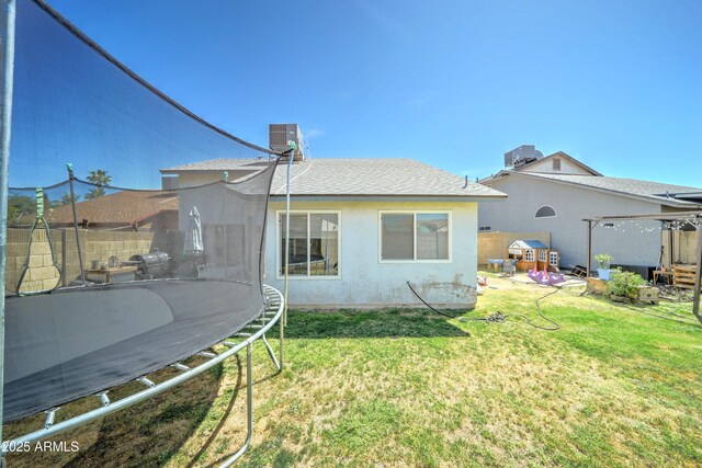 rear view of house with a trampoline, a shingled roof, a yard, and central AC unit