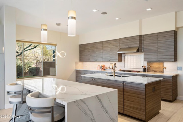 kitchen with modern cabinets, an island with sink, under cabinet range hood, a sink, and decorative backsplash