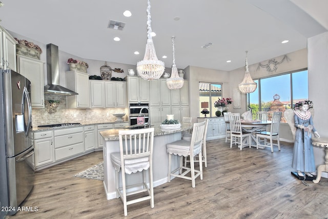 kitchen featuring a kitchen island with sink, light hardwood / wood-style flooring, light stone counters, wall chimney exhaust hood, and appliances with stainless steel finishes