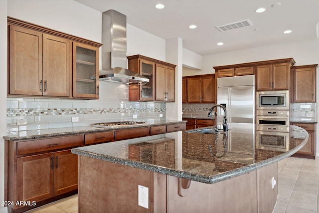 kitchen with built in appliances, a center island with sink, wall chimney exhaust hood, and sink