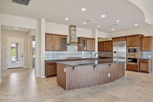 kitchen with built in appliances, dark stone counters, a breakfast bar area, wall chimney range hood, and a large island